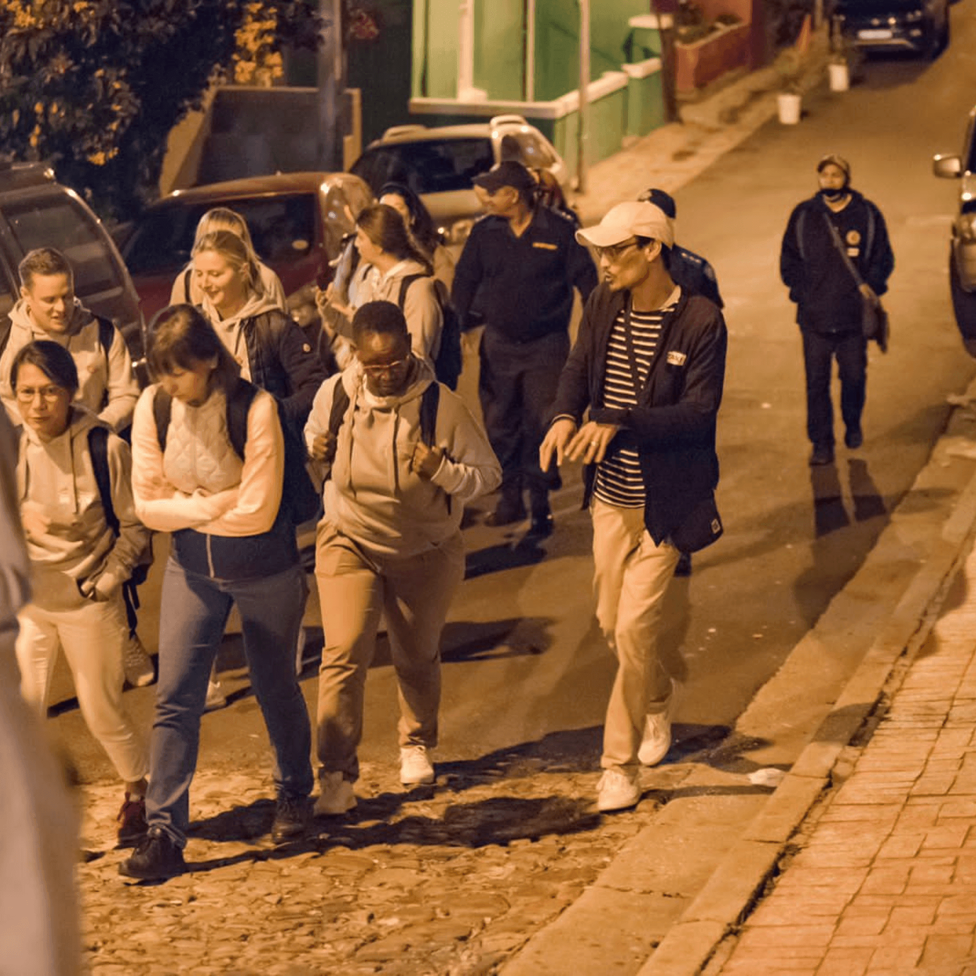 Group of people walking up a cobblestone street at night, some holding backpacks.